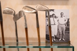 Vintage hickory golf clubs displayed on a glass shelf, with a historic black-and-white photograph of early 20th-century golfers in the background, evoking the material history and tradition of the game