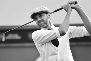Paolo Quirici, dressed in vintage golf attire with a flat cap and tie, follows through on a golf swing using a hickory club, captured in black and white