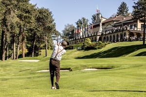 Golfer playing an approach shot with a hickory golf club toward the green in front of the Villa d’Este clubhouse.