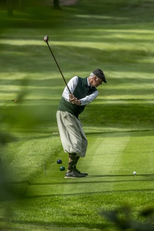Golfer in vintage attire completing a swing with a hickory golf club on a tree-lined fairway during the Hickory Masters.
