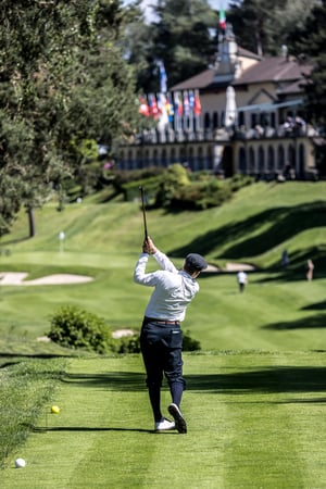 Golfer teeing off with a hickory club while spectators watch from the hillside beside the Villa d’Este clubhouse.
