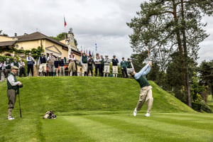 Golfer teeing off with a hickory club while spectators watch from the hillside beside the Villa d’Este clubhouse.