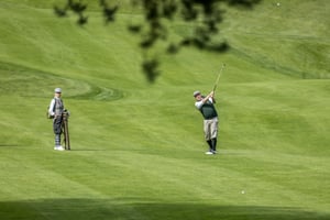 Golfer striking a shot with a vintage hickory club as another participant carries the traditional golf bag on the fairway.