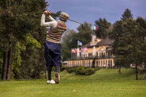 Hickory golfer completing a tee shot with the Villa d’Este clubhouse and national flags in the background.