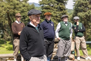 Group of hickory golfers in period attire listening attentively during the tournament briefing.