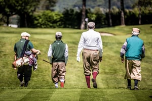 Four golfers dressed in traditional hickory golf attire walk together down a lush fairway carrying vintage golf clubs during a practice round.