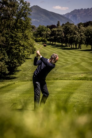 Golfer swings a vintage wooden driver from the tee with a panoramic mountain backdrop overlooking the fairway.