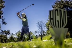 Low-angle view of a hickory golfer teeing off with a vintage wooden driver beside the Hickory Golf Masters tee marker.