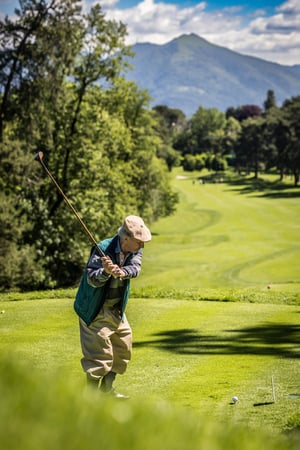 Golfer in traditional hickory golf clothing begins a tee shot with a vintage wooden club against a mountain landscape.