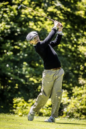 Low-angle view of a hickory golfer at the top of the backswing beside an HGM tee marker before driving from the tee box.