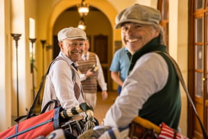 Two smiling hickory golfers carrying vintage golf bags arrive inside the clubhouse before the practice round.
