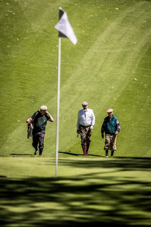Three golfers in traditional hickory golf attire walk up the fairway toward the green during a practice round.