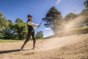 Golfer wearing vintage hickory golf apparel plays a bunker shot as sand sprays into the air under a clear blue sky.