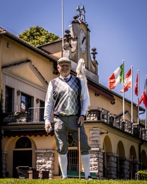 Hickory golfer in vintage golf clothing stands smiling in front of the historic clubhouse holding a wooden golf club.