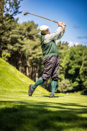 Golfer in traditional hickory golf attire completes a full follow-through with a vintage wooden driver on the fairway.