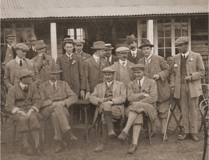 Group of early 20th-century golfers and officials gathered outside a clubhouse, dressed in traditional tweed jackets, flat caps, and ties, some seated and others standing, posing formally for a group photograph.
