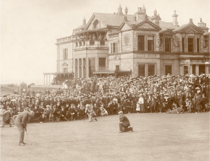 Historic golf scene at St Andrews with a large crowd gathered around the green, watching a golfer putt in front of the iconic Royal and Ancient Clubhouse.