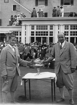 Two men in vintage golf attire holding a large ornate trophy together, standing in front of a clubhouse with a crowd of spectators gathered behind them and on the rooftop.