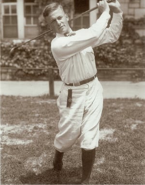 Young Bobby Jones captured mid-follow-through in a classic golf pose, dressed in early 20th-century attire with high socks, plus fours, and a bow tie, standing on grass in front of a building.