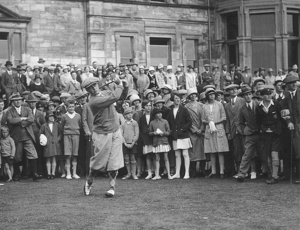 Bobby Jones mid-swing during the 1927 British Open, surrounded by a large crowd of spectators in period attire in front of the Royal and Ancient Clubhouse at St Andrews.