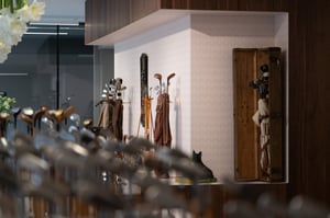 Soft-focus view of antique golf clubs in the foreground with vintage golf bags and memorabilia displayed against patterned walls.