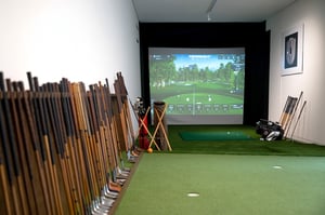 Golf simulator room featuring a projected virtual fairway, surrounded by rows of vintage hickory-shaft golf clubs along the walls.