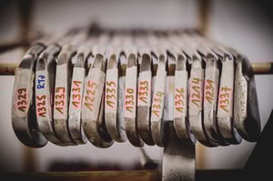 Close-up of vintage iron clubheads, each labeled with handwritten inventory numbers on masking tape.