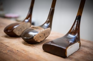 Close-up of three wooden-head golf clubs on a workbench, showing restoration patches and polished wooden grain.