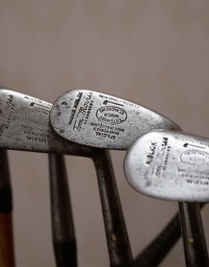 Close-up of vintage hickory-shaft golf irons resting on a wooden surface, showing worn steel heads and traditional whipping around the hosels.