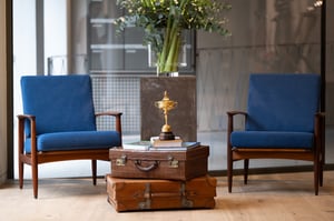 Two blue mid-century chairs facing a small table made of stacked vintage suitcases, topped with a gold golf trophy and books.