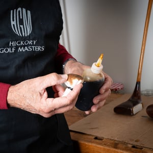 Close-up of an artisan applying wood stain to the head of a vintage hickory golf club, with the Hickory Golf Masters logo visible on the apron.