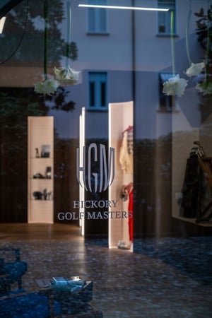 Reflection on a glass window with the Hickory Golf Masters logo, revealing illuminated interior displays featuring vintage golf accessories.