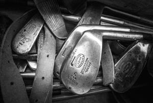 Close-up view of antique hickory-shafted golf irons, featuring engraved markings from T. Stewart and other historic makers, lying together with leather straps